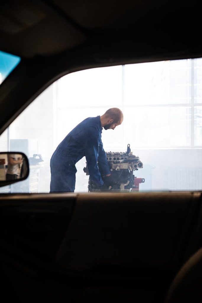 Mechanic working on an engine inside a garage, viewed through a car window.